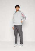 Junger Mann, der einen hellgrauen Hoodie mit rotem Schriftzug auf dem Ärmel, dunkelgraue Hose und weiße Sneaker trägt, mit einer Hand in der Tasche stehend.