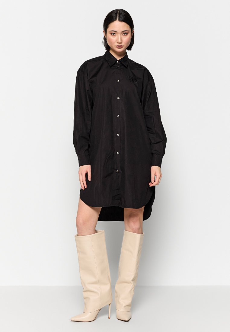 Schwarzes, übergroßes Hemdkleid mit langen Ärmeln, Knopfverschluss und spitzem Kragen. Getragen mit knielangen beigen Stiefeln. Glatte Textur.