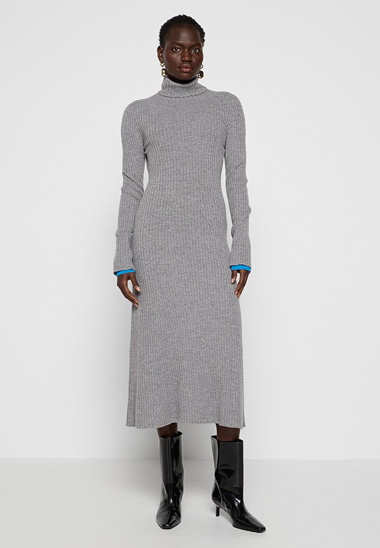 Frau in einem grauen, gerippten Midi-Kleid mit Rollkragen, schwarzen, hochhackigen Stiefeln und goldenen Creolen, die vor einem weißen Hintergrund steht.