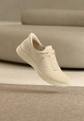 Hellbeiger gestrickter Sneaker mit Schnürsenkeln, perforiertem Design und flexiblem Sohlen, schwebend über beige Teppich in einem Raum mit passender Einrichtung.