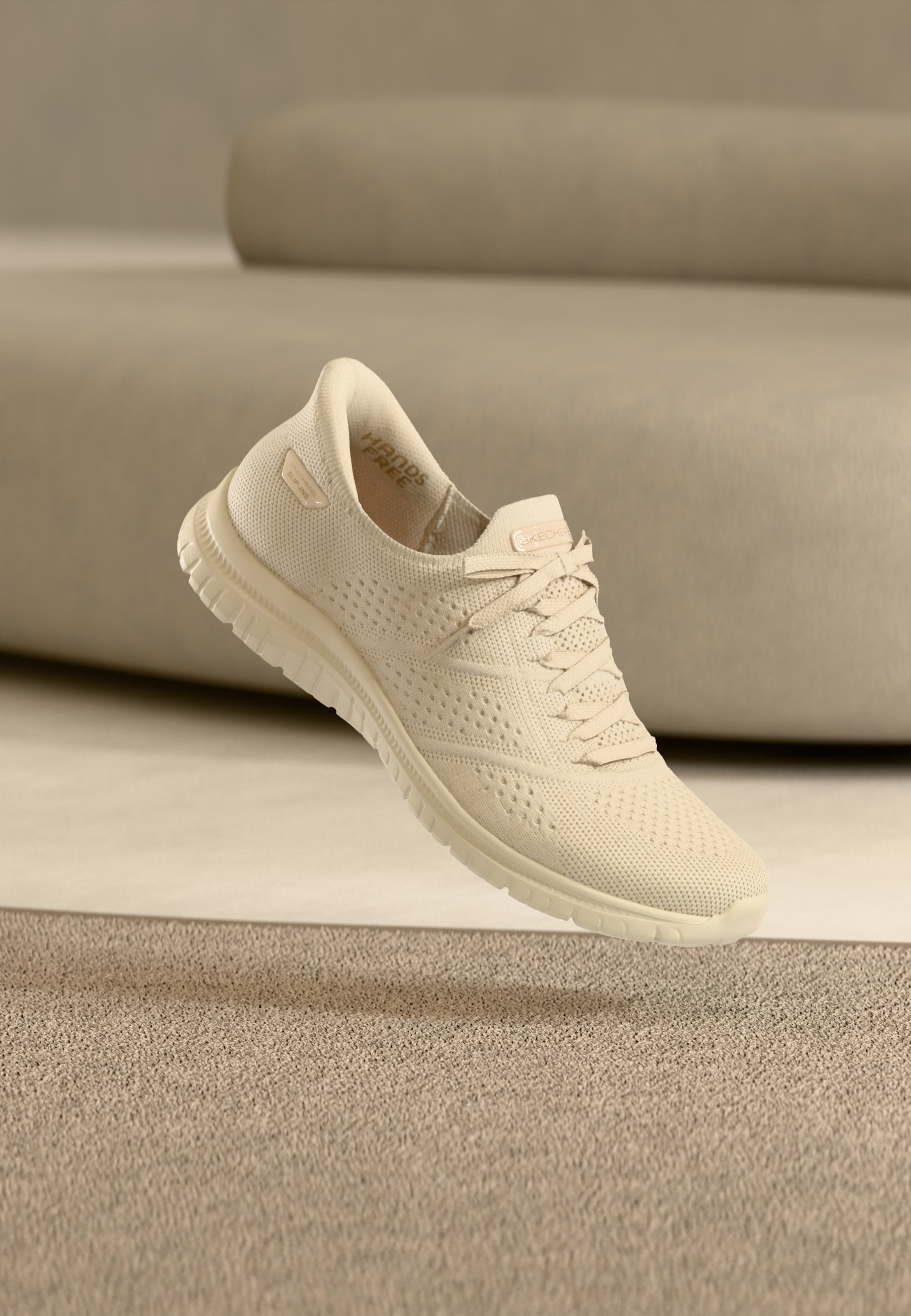 Hellbeiger gestrickter Sneaker mit Schnürsenkeln, perforiertem Design und flexiblem Sohlen, schwebend über beige Teppich in einem Raum mit passender Einrichtung.