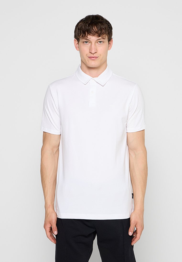 Junger Mann, der nach vorne blickt, trägt ein schlichtes weißes kurzärmeliges Poloshirt und schwarze Hose vor neutralem Hintergrund.