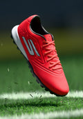 Leuchtend roter Fußballschuh mit schwarzer Sohle, geschnürte Schnürsenkel, schwebt über einem Grasfeld mit weißer Begrenzungslinie und fliegenden Grasblättern.