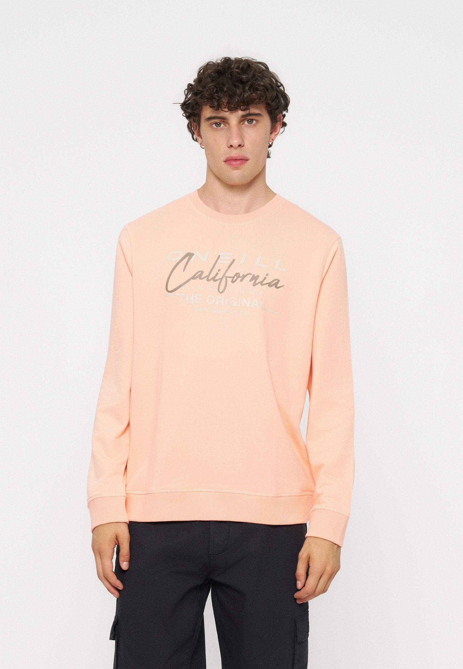 Junger Mann mit lockigem dunklem Haar, der einen hellen pfirsichfarbenen Sweatshirt mit dem Schriftzug „California“ und dunkle Hosen trägt, steht vor einem schlichten weißen Hintergrund.