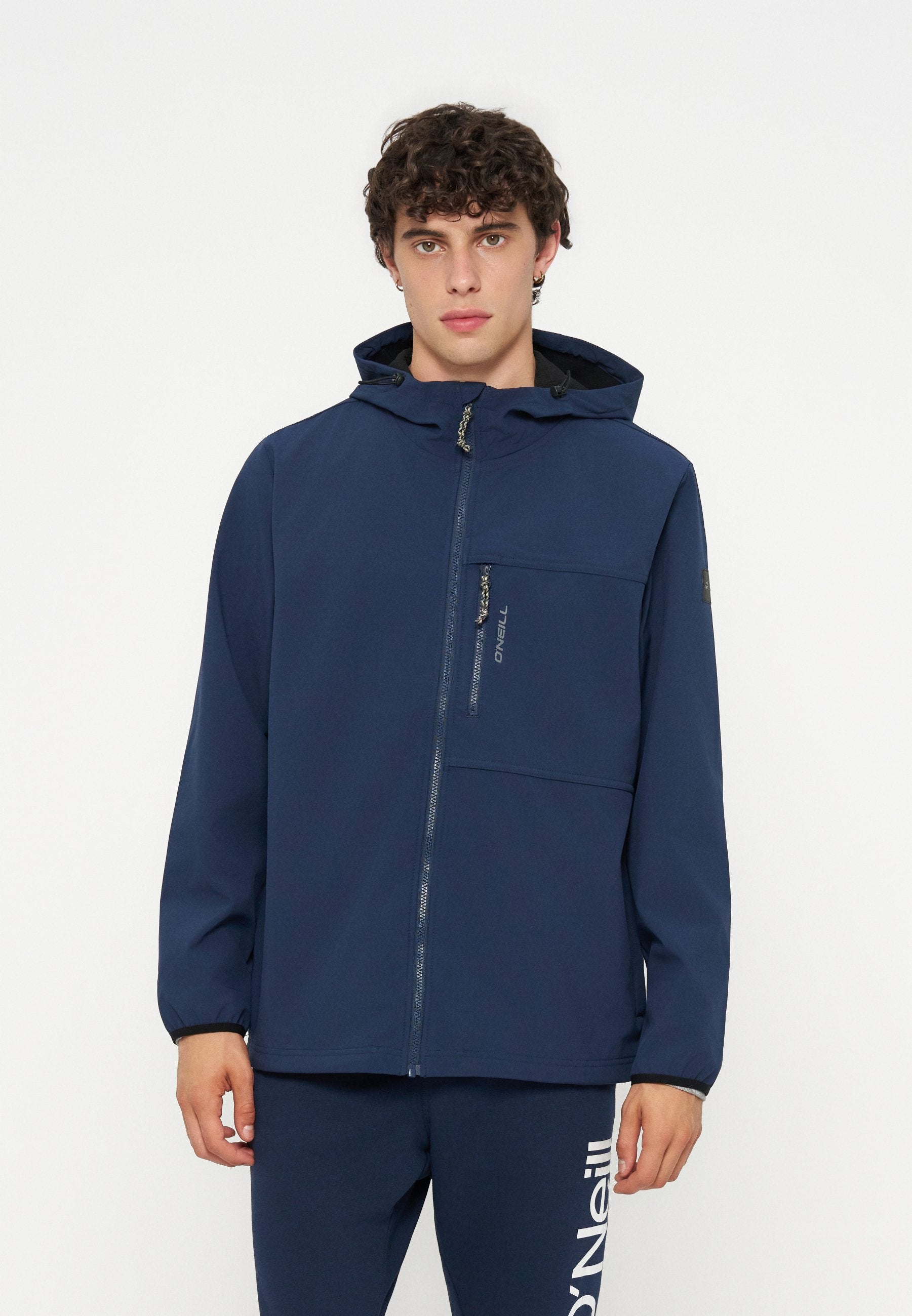 Junger Mann mit lockigem Haar, der eine marineblaue Kapuzenjacke und passende Hose mit O’Neill-Logo trägt, steht vor weißem Hintergrund.