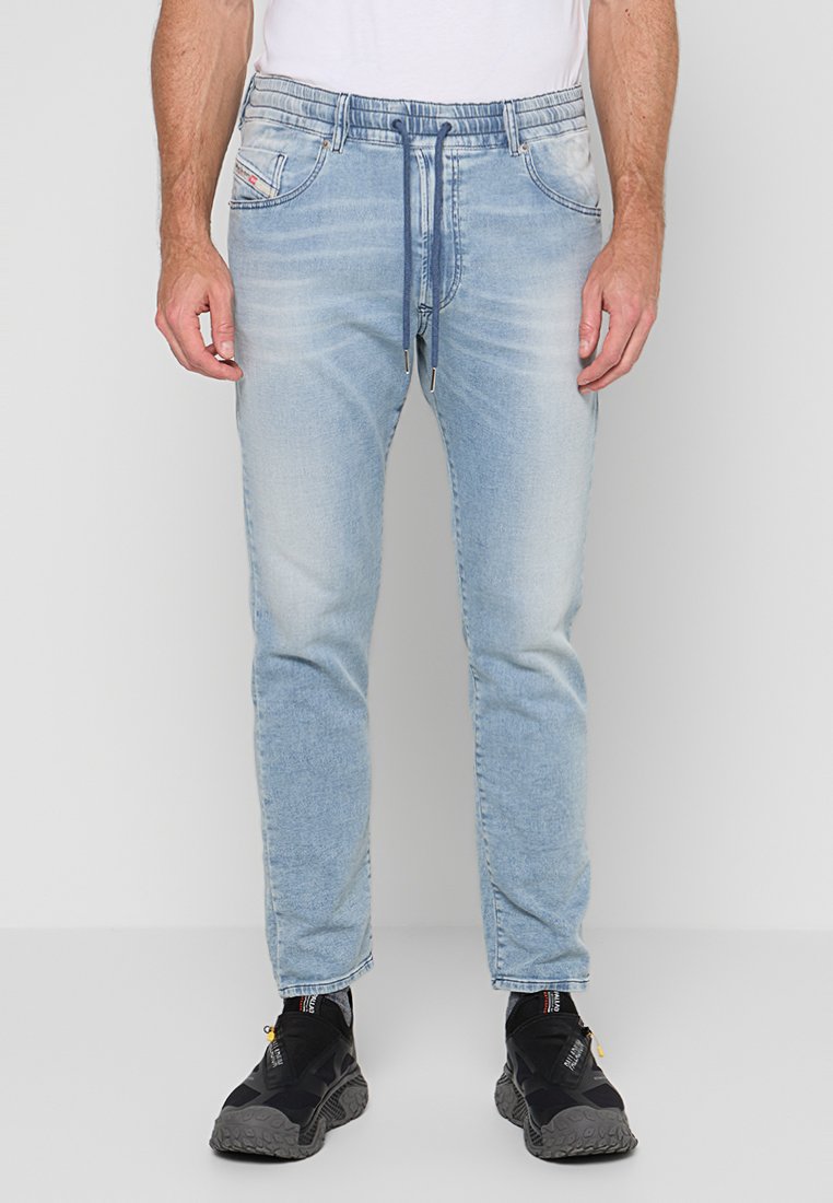 Mann trägt hellblaue Denim-Jogger-Jeans mit verstellbarem Bund und schwarze Sportschuhe und steht vor einem weißen Hintergrund.