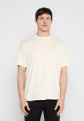 Junger Mann mit kurzem braunem Haar, der ein schlichtes, cremefarbenes T-Shirt mit kurzen Ärmeln und schwarze Hosen trägt, vor einem einfarbigen Hintergrund steht.
