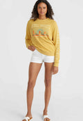 Gelbes Sweatshirt mit Grafikdruck, lange Ärmel, gerippte Bündchen, kombiniert mit weißen Shorts. Modell trägt offene Sandalen und steht vor einem schlichten Hintergrund.