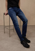 Dunkelblaue Denim-Jeans mit schmaler Passform, detaillierter Nahtführung und Seitentaschen, kombiniert mit schwarzen texturierten Schuhen, sitzend auf einem Metallhocker.
