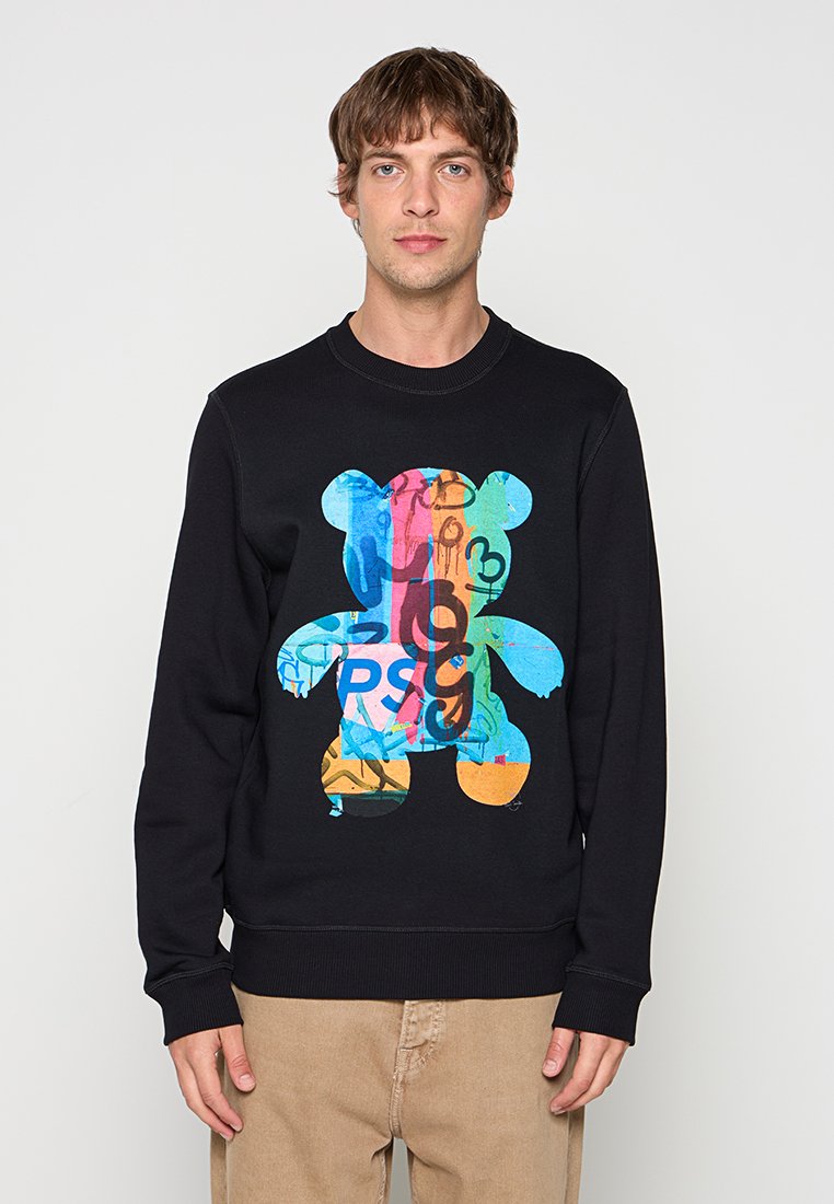 Junger Mann trägt schwarzen Sweatshirt mit großem buntem Graffiti-Bären auf der Vorderseite in der Mitte, kombiniert mit beigen Hosen.