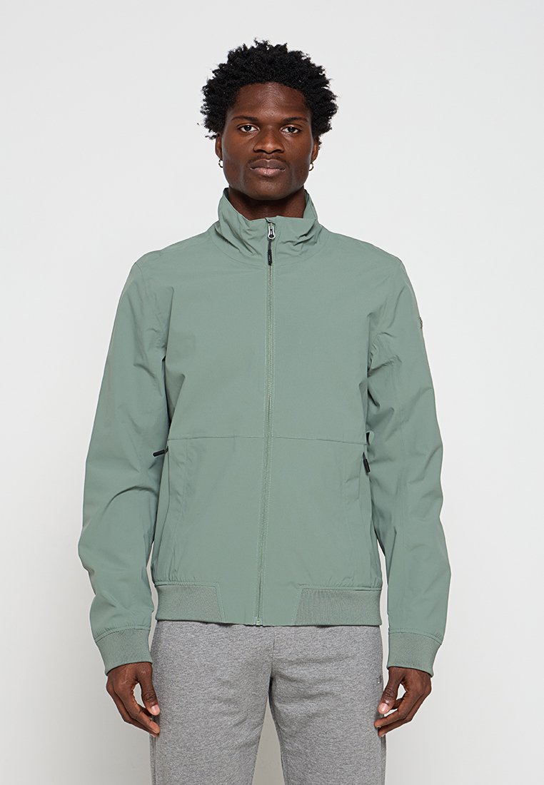 Mann trägt hellgrüne Reißverschlussjacke und graue Jogginghose, steht vor einem einfarbigen Hintergrund und schaut geradeaus.