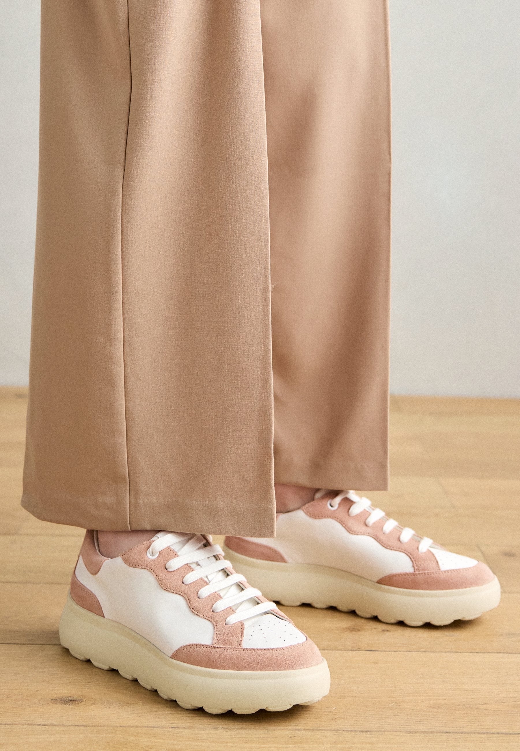 Weiße Sneaker mit rosa Wildleder-Akzenten, runder Zehenform, dicker cremefarbener Sohle und Schnürdesign. Getragen mit beigen, weit geschnittenen Hosen.