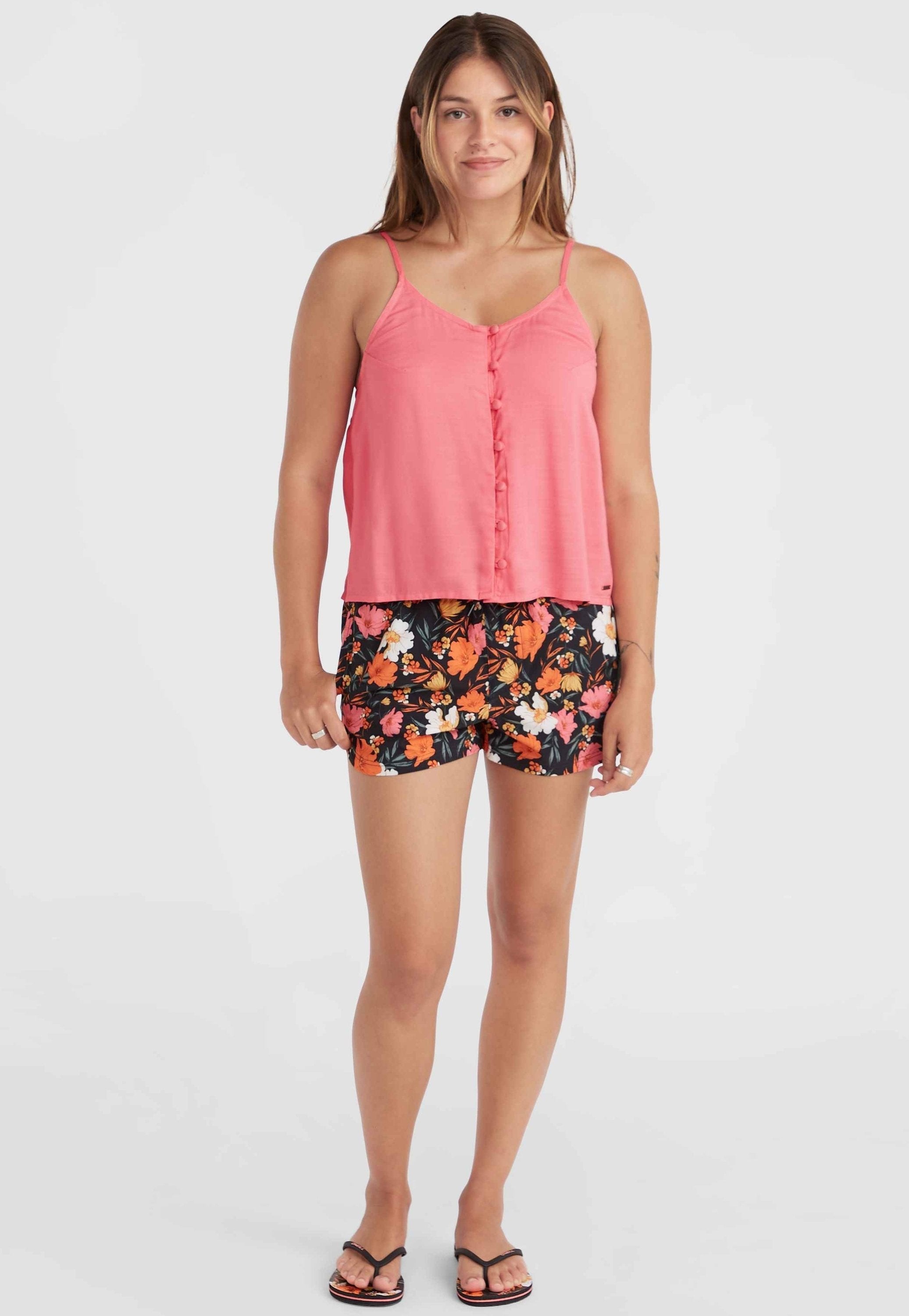 Rosa ärmelloses Top mit vorderen Knöpfen, kombiniert mit schwarzen Shorts mit einem bunten Blumenmuster. Das Model trägt schwarze Sandalen.
