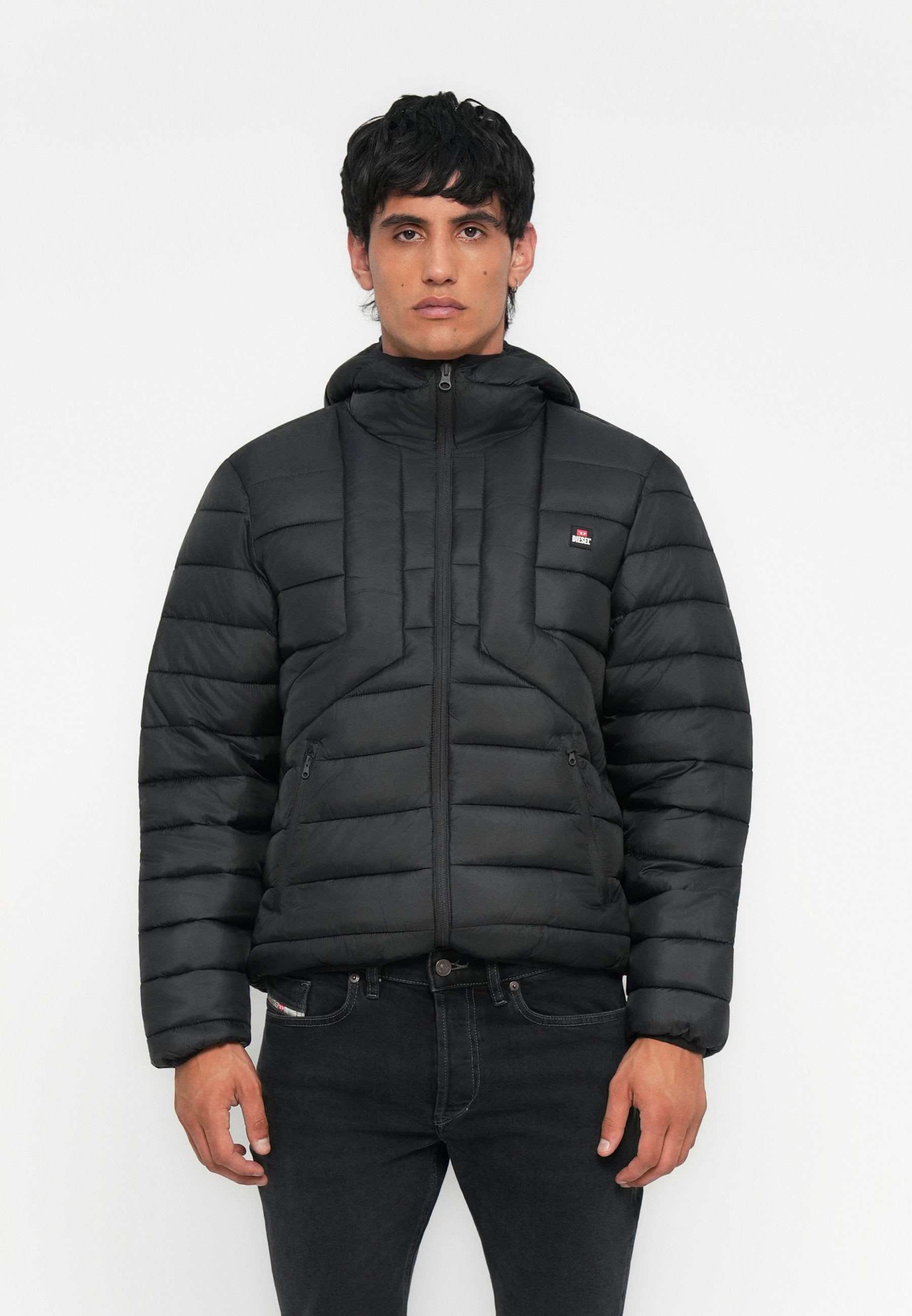Mann in einer schwarzen gefütterten Jacke mit Kapuze, der aufrecht steht und nach vorne schaut, vor einem schlichten weißen Hintergrund.