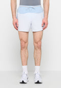 Mann trägt hellgraue Sportshorts mit einem blauen Bund, weiße Socken und weiße Laufschuhe, steht vor einem schlichten Hintergrund.