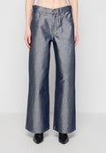 Frau trägt hoch taillierte, weit geschnittene metallicblaue Jeans und schwarze spitze Stiefel, steht vor einem schlichten hellen Hintergrund.
