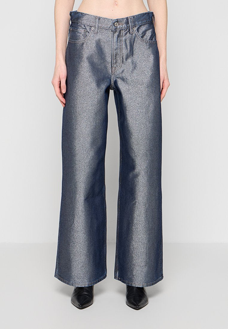 Frau trägt hoch taillierte, weit geschnittene metallicblaue Jeans und schwarze spitze Stiefel, steht vor einem schlichten hellen Hintergrund.