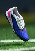Athletischer Schuh mit einem strukturierten weißen und blauen Obermaterial, schwarzen Akzenten, pinkem Swoosh und einer gemusterten Zehenkappe mit multidirektionalen Noppen.