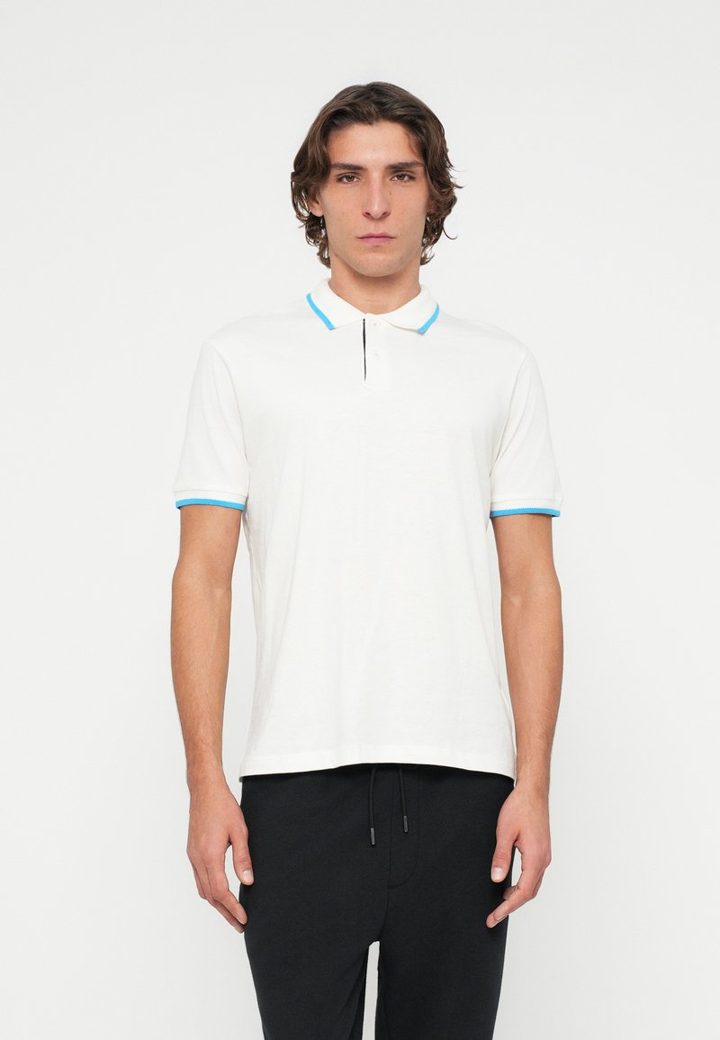 Junger Mann, der ein weißes Poloshirt mit blauem Besatz und schwarze Hose trägt, vor einem einfachen weißen Hintergrund stehender.