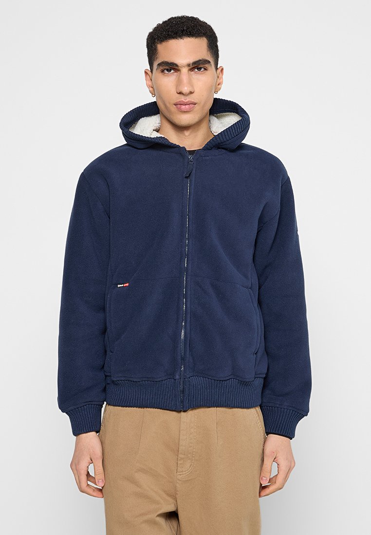 Marineblaue Fleece-Hoodie mit Reißverschluss vorne, gerippten Bündchen und Saum. Verfügt über eine gefütterte Kapuze und einen kleinen Logoakzent auf der Taschenseite.