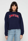 Frau mit dunklen Haaren, die einen marineblauen Pullover mit dem Schriftzug "LOVER" in roten Buchstaben und einem roten Herz trägt, kombiniert mit hellblauen Jeans.