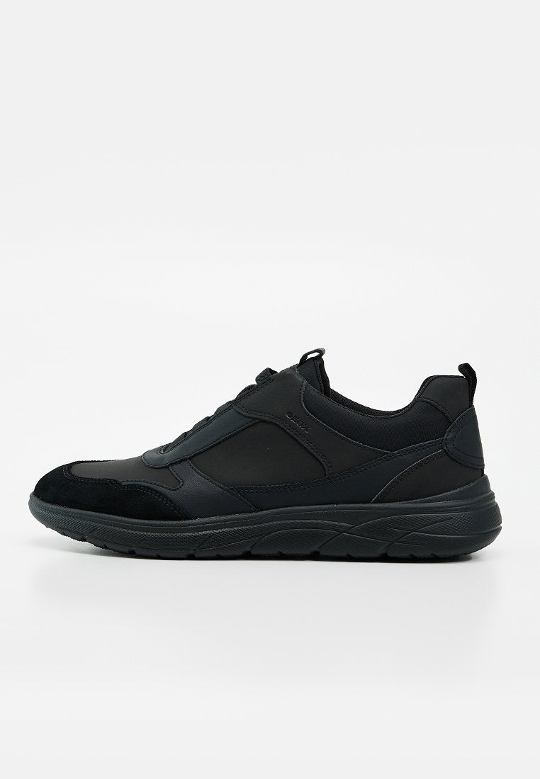 Schwarzer Low-Top-Sneaker mit strukturierter Sohle, gemischten Leder- und Stoffpaneelen sowie Zuglaschen an Zunge und Ferse vor weißem Hintergrund.
