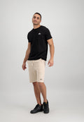 Schwarzes T-Shirt mit kurzen Ärmeln und kleinem Logo auf der Brust, kombiniert mit beigen Shorts mit Kordelzug und schwarzen Sportschuhen. Schlichter Hintergrund.