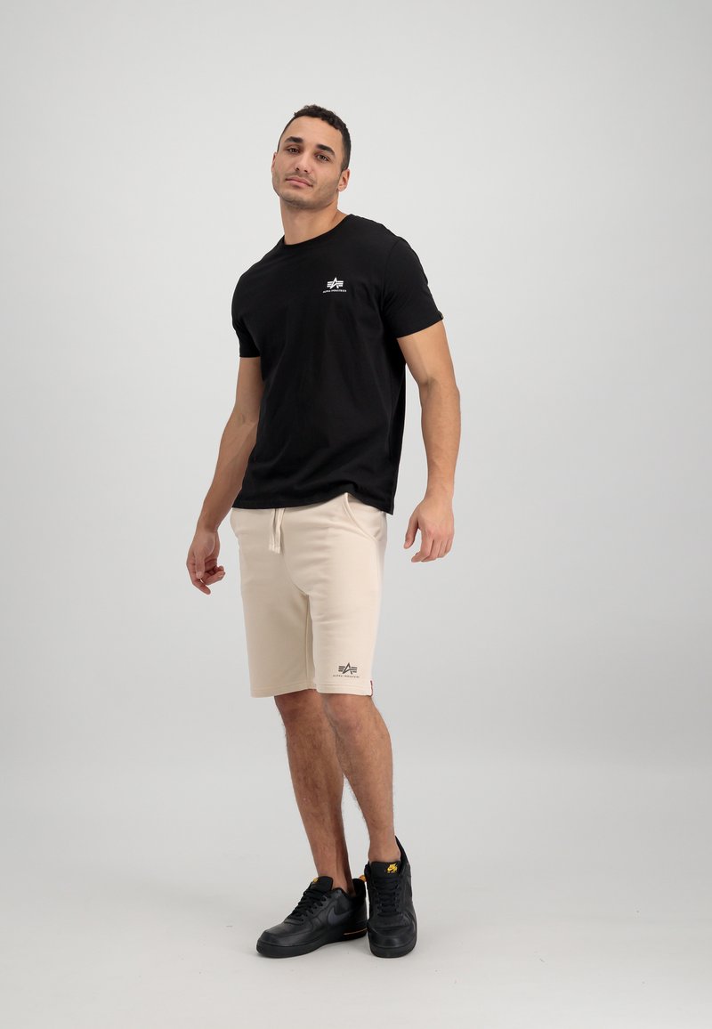 Schwarzes T-Shirt mit kurzen Ärmeln und kleinem Logo auf der Brust, kombiniert mit beigen Shorts mit Kordelzug und schwarzen Sportschuhen. Schlichter Hintergrund.