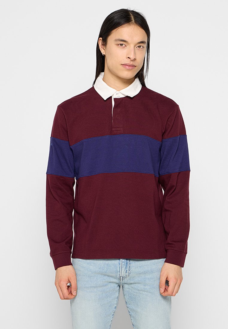 Junge Person mit langen schwarzen Haaren, die ein burgunderfarbenes und navyblaues Rugbyhemd mit weißem Kragen und hellblauen Jeans trägt, steht vor einem schlichten Hintergrund.