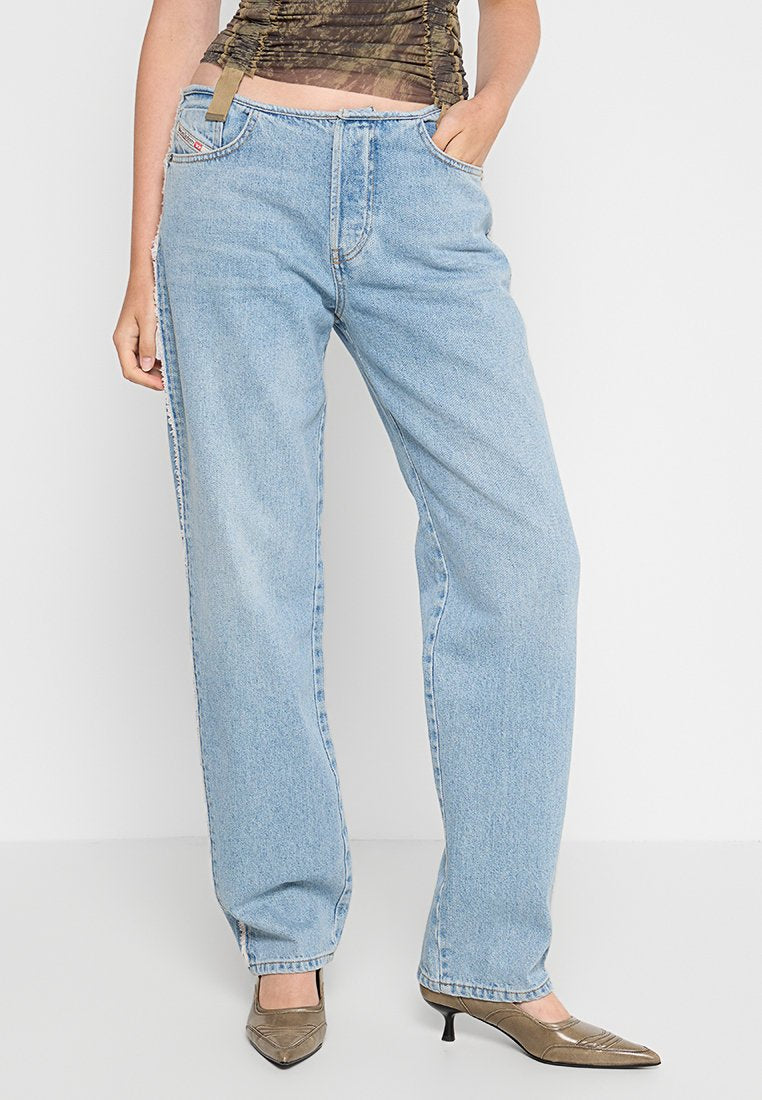 Frau trägt hochtaillierte, hellblaue, gerade geschnittene Jeans, die Hände in den Taschen, kombiniert mit braunen, spitzen Kitten-Heel-Schuhen und einem gerafften Oberteil.