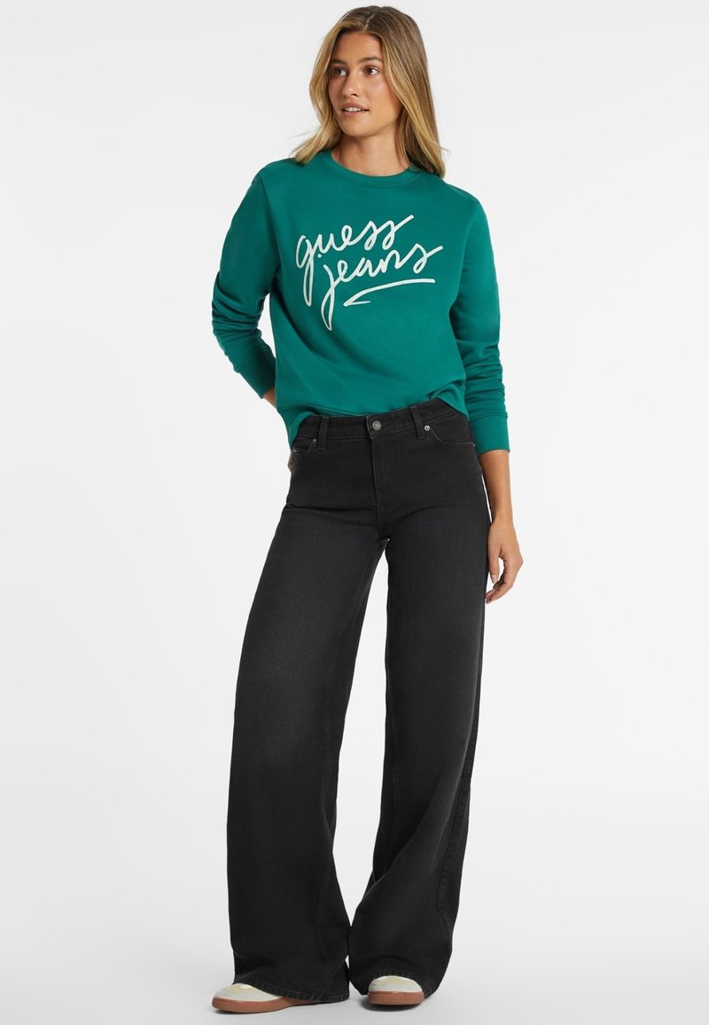 Türkiser Sweatshirt mit "guess jeans" in weißer Schrift, kombiniert mit schwarzen weiten Jeans. Das Outfit zeichnet sich durch einen lässigen, lockeren Schnitt aus.