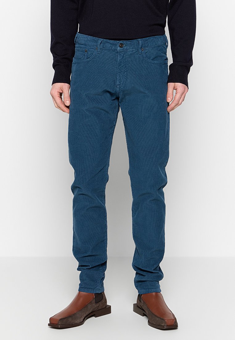 Mann in blauen Cordhosen, schwarzem Langarmoberteil und braunen Leder-Schlüpfschuhen, der vor einem weißen Hintergrund steht.
