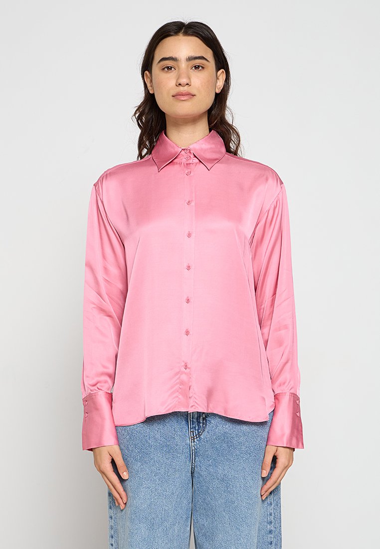 Rosa Satin-Hemd mit langen Ärmeln, Kragen, vorderen Knöpfen und Manschettendetails. Glatte Textur und lässige Passform. Getragen mit hellblauen Jeans.