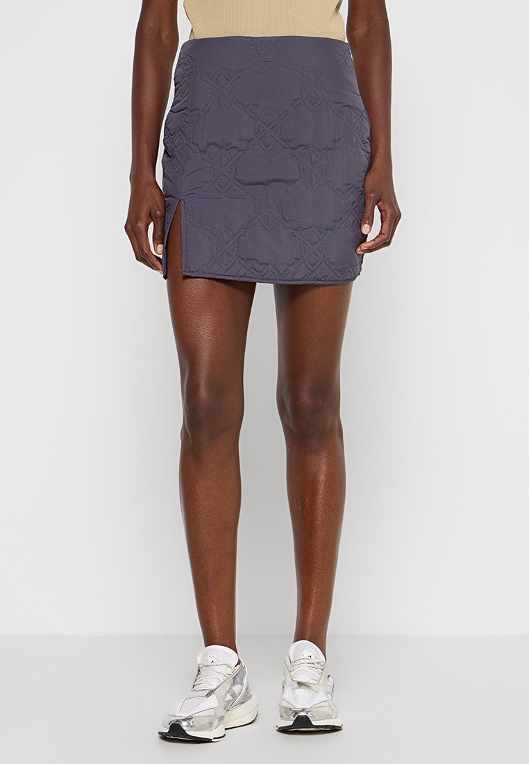 Frau in einem marineblauen, gesteppten Minirock mit seitlichem Schlitz und weißen Sneakers, stehend vor einem einfarbigen Hintergrund.