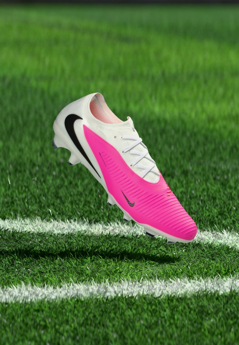 Weißer und leuchtend pinker Nike-Fußballschuh mit schwarzem Swoosh, schwebend über grünem Gras nahe der weißen Spielfeldlinie.