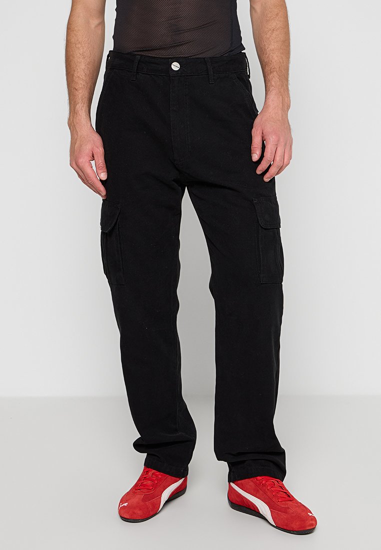 Schwarze Cargo-Hosen mit mehreren Taschen, aus strapazierfähigem Stoff, mit geradem Bein und Knopfverschluss am Bund. Rote Schuhe ergänzen den Look.