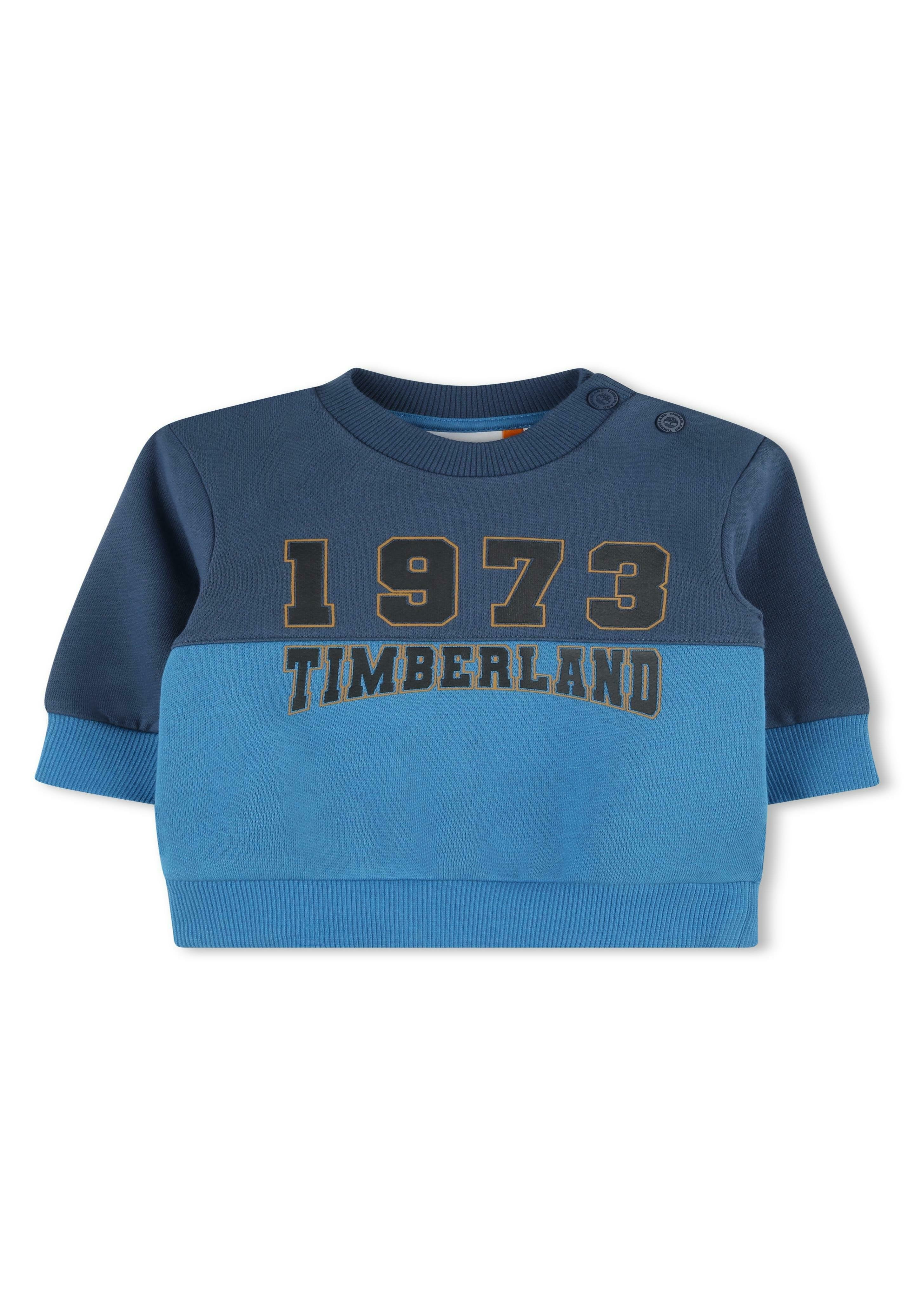 Blaues und dunkelblaues Sweatshirt mit langen Ärmeln, gerippten Bündchen, rundem Halsauschnitt und "1973 TIMBERLAND" in Gold gedruckt. Zwei Druckknöpfe an der Schulter.