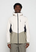 Mann trägt eine Kapuzenjacke im Colorblock-Design mit weißem Oberteil, grauem Unterteil und schwarzen Seitenpartien, steht vor einem schlichten grauen Hintergrund.