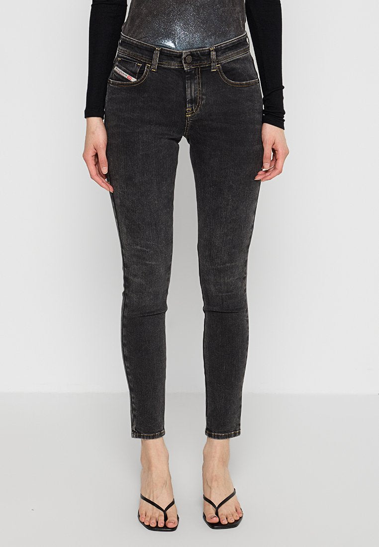 Schwarze Skinny-Jeans mit einer abgedunkelten Textur, ausgestattet mit fünf Taschen und gelben Nähten. Kombiniert mit einem eng anliegenden schwarzen Oberteil und minimalistischen Sandalen.