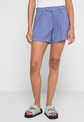 Frau in blauen, taillierten High-Waist-Shorts mit Knopf, weißem Oberteil und hellblauen Doppelsandaletten vor weißem Hintergrund.