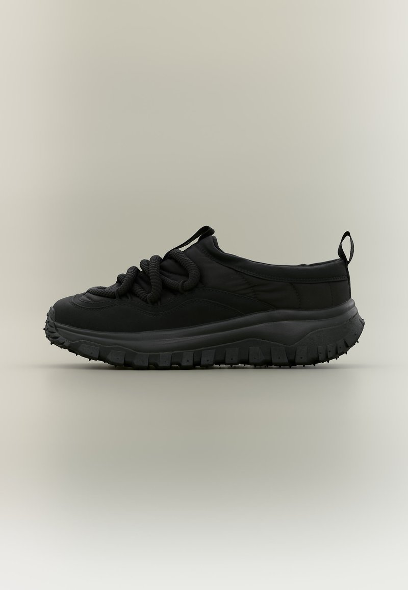 Schwarzer Sneaker mit niedrigem Schaft, dicker strukturierter Sohle und groben Schnürsenkeln im Kordelstil vor neutralem Hintergrund, Seitenansicht.