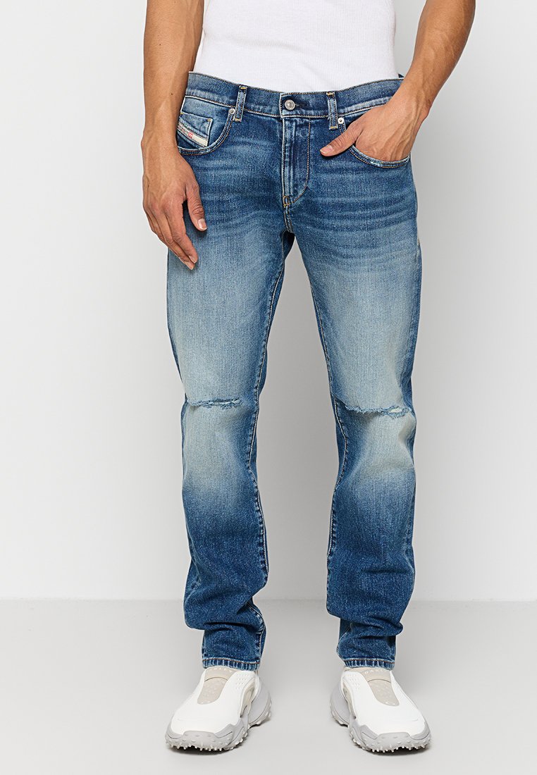 Mann trägt ausgebleichte blaue Jeans mit leichten Abnutzungserscheinungen an den Knien, weiße Slip-On Sneakers und ein weißes ärmelloses Shirt.