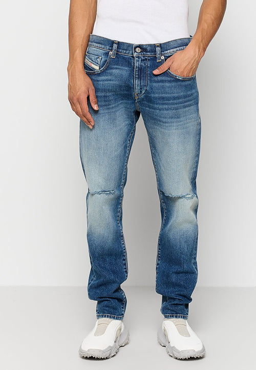 Mann trägt ausgebleichte blaue Jeans mit leichten Abnutzungserscheinungen an den Knien, weiße Slip-On Sneakers und ein weißes ärmelloses Shirt.