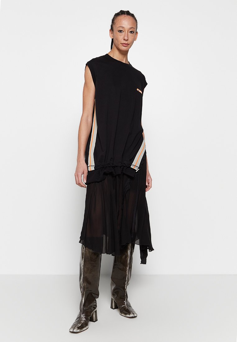 Schwarzes ärmelloses Kleid mit taillierter Taille, geschichtetem transparentem Rock und seitlichen Streifen in Orange, Weiß und Grau. Modell trägt metallische Stiefel.