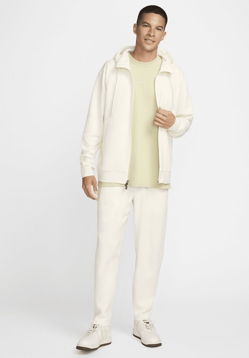 Weiße Zip-Hoodie über einem hellgrünen T-Shirt, kombiniert mit weißen Hosen und weißen Turnschuhen. Glatter Stoff, lockere Passform, einfaches Design.