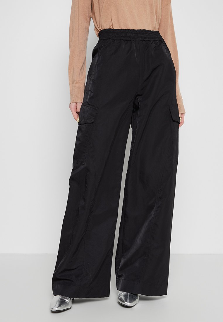 Schwarze Cargo-Hosen mit lockerer Passform, elastischem Bund, zwei Seitentaschen und einer satinartigen Textur, kombiniert mit einem beigen Oberteil und silbernen Schuhen.