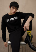 Schwarzer Pullover mit der Aufschrift "DIESEL", kombiniert mit schwarzen Hosen. Das Modell trägt gelbe Socken mit schwarzen Akzenten und schwarze Stiefel mit Reißverschluss.
