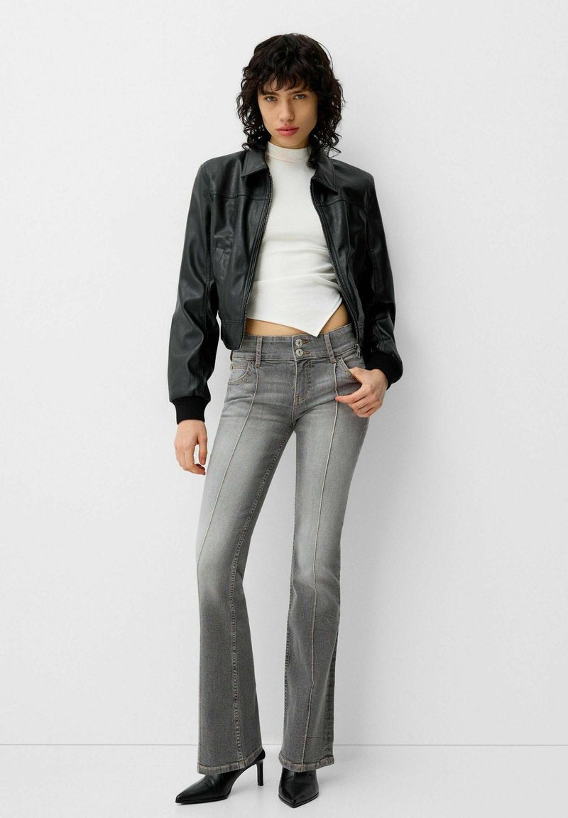 Schwarze Leder-Bomberjacke über einem weißen, cropped Top, kombiniert mit grauen Schlagjeans mit Kontrastnähten und einer Drei-Knopf-Verschluss.