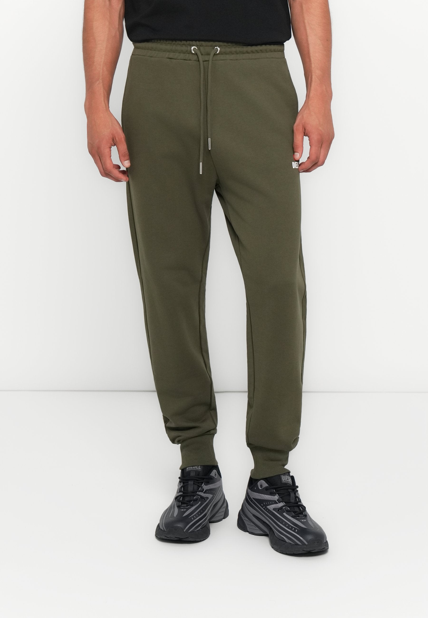 Olivgrüne Sweatpants mit elastischem Bund, verstellbarem Kordelzug und seitlichen Streifen. Kombiniert mit schwarzen Sportschuhen.