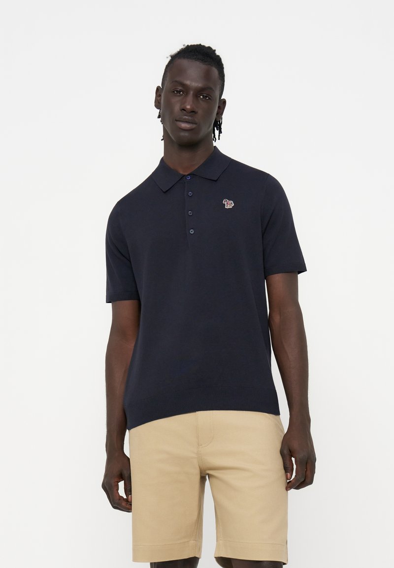 Mann trägt ein schwarzes kurzärmliges Polo mit kleinem Zebra-Logo und beigefarbenen knielangen Shorts, steht vor einem weißen Hintergrund.